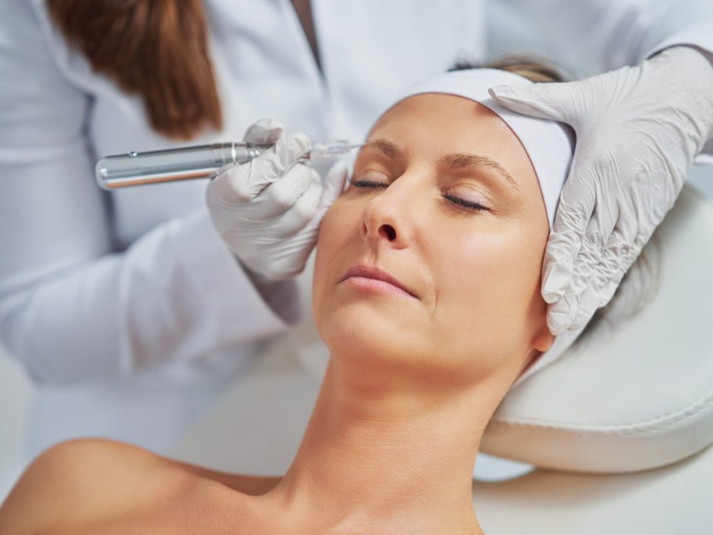Micropigmentación de cejas con dermógrafo: una profesional con guantes blancos trabaja la ceja de una clienta tumbada con diadema y los ojos cerrados, cejas tatuadas Zaragoza